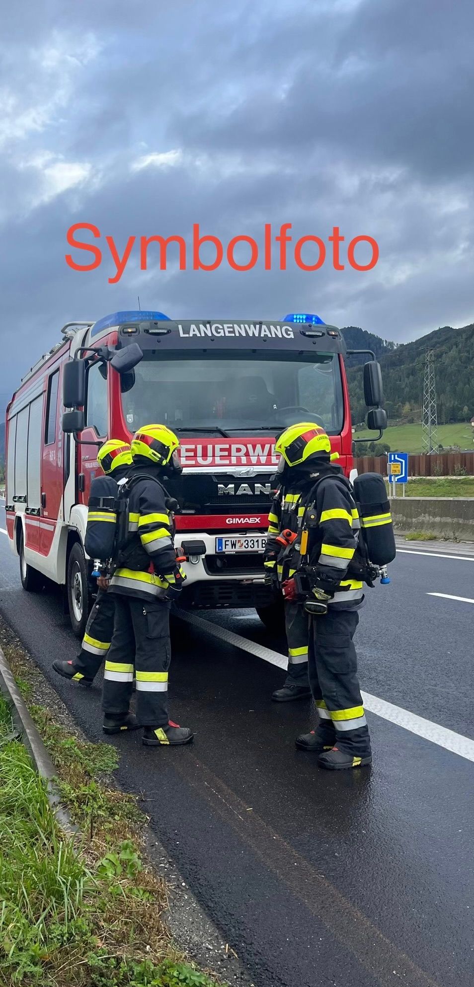 Three firefighters in full gear stand beside a red fire truck with the word 'EUERWE' on the front, parked on the side of a road. The sky is cloudy.