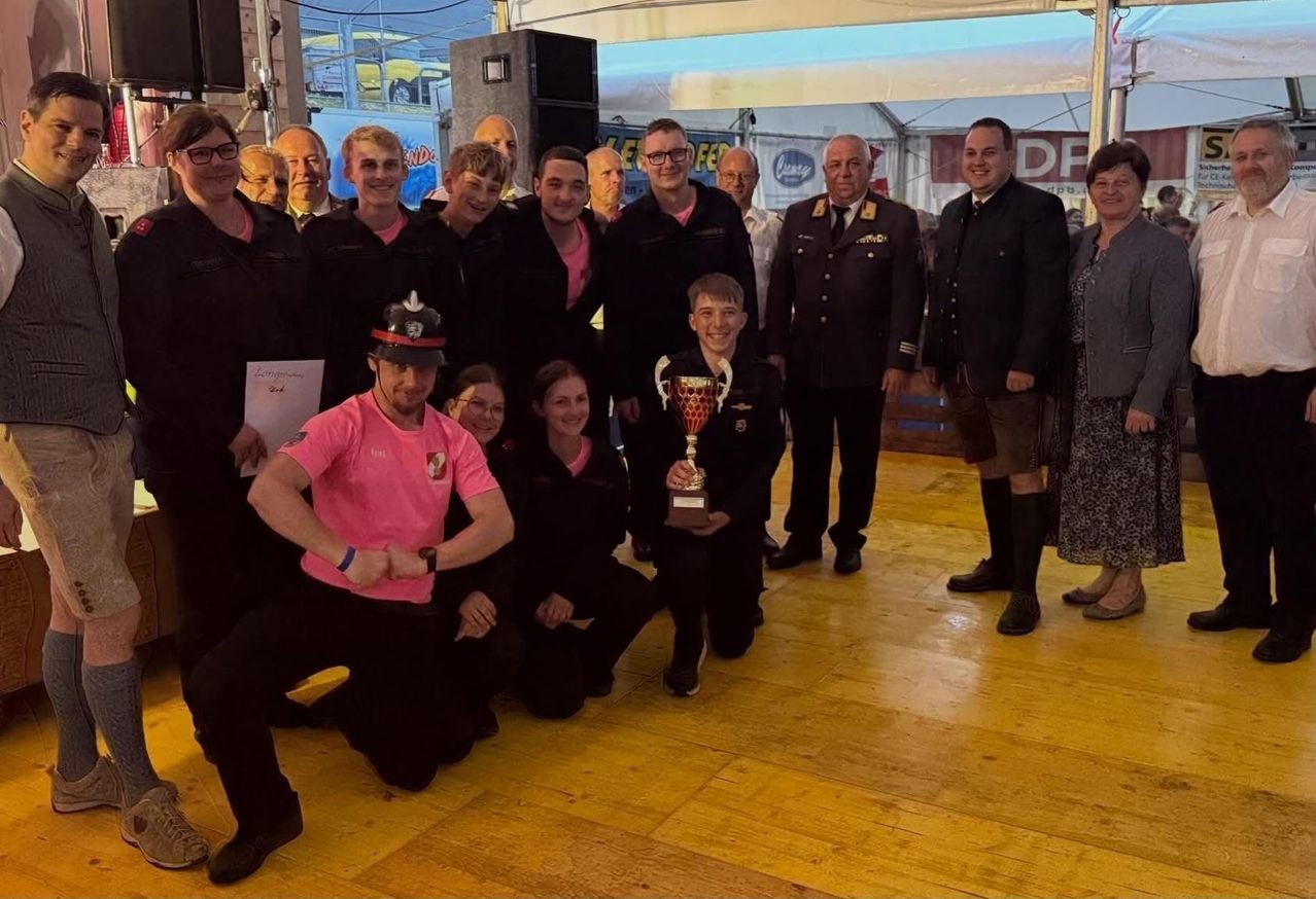 Eine Gruppe von Menschen in Uniform und Zivilkleidung posiert für ein Foto mit einer Trophäe. Einige knien, andere stehen und manche tragen Hüte. Sie befinden sich bei einer Indoor-Veranstaltung mit Holzboden.