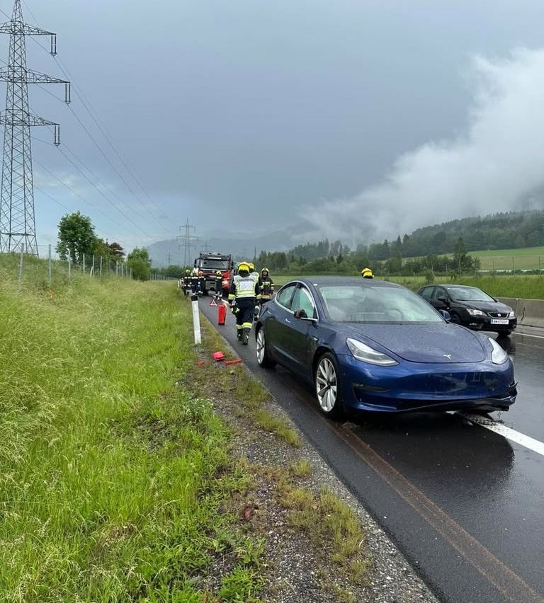 Emergency services are attending to a blue Tesla Model 3 on a wet road. Firefighters in yellow helmets and reflective vests are near the car.