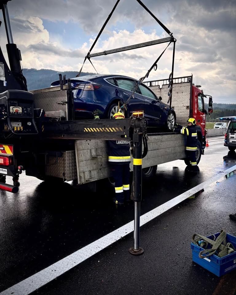A blue Tesla Model 3 is being lifted onto a tow truck. Two workers in safety gear are standing by.