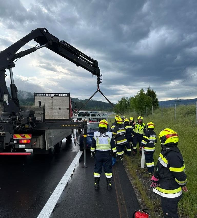 Firefighters in protective gear stand on the roadside with a wrecked car. A crane is lifting a damaged car. The sky is cloudy.