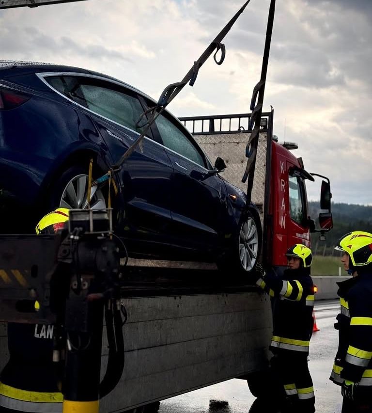 Firefighters are removing a blue car from a flatbed truck. The car is held by chains and is sitting on a grey surface. The firefighters wear yellow helmets.