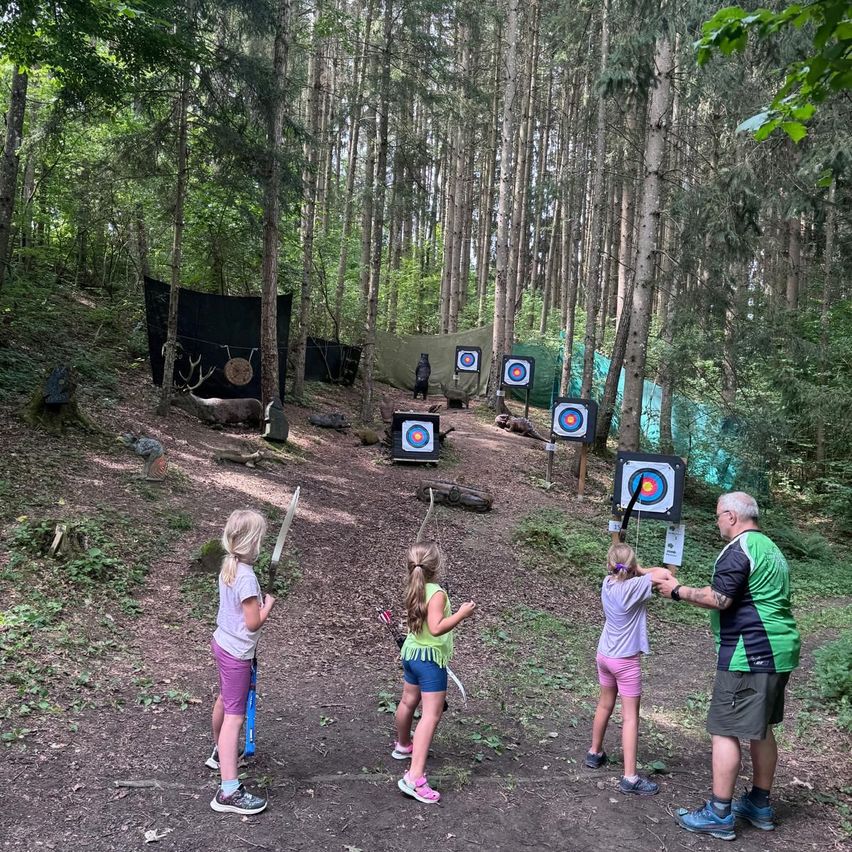 Vier Kinder und ein erwachsener Mann üben Bogenschießen im Wald. Die Kinder halten Bögen und Pfeile, und der Mann gibt ihnen Anweisungen. Der Wald ist voller Bäume, und es gibt mehrere Ziele auf dem Boden aufgestellt.