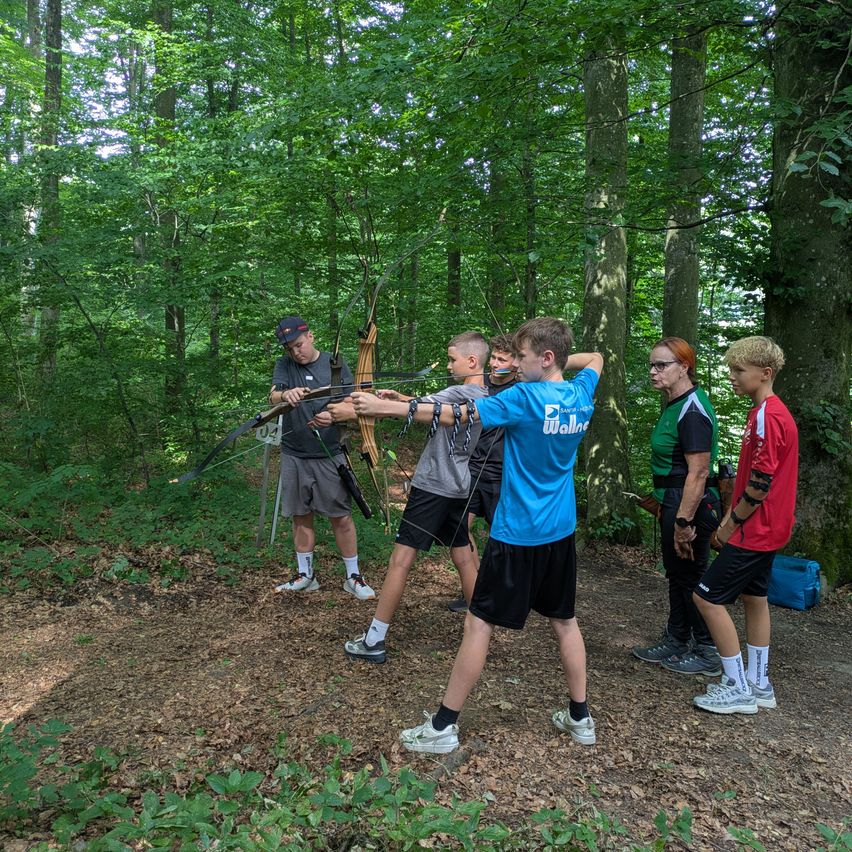 Eine Gruppe junger Menschen und Erwachsener übt Bogenschießen im Wald. Der Junge im blauen Shirt zielt mit seinem Bogen.