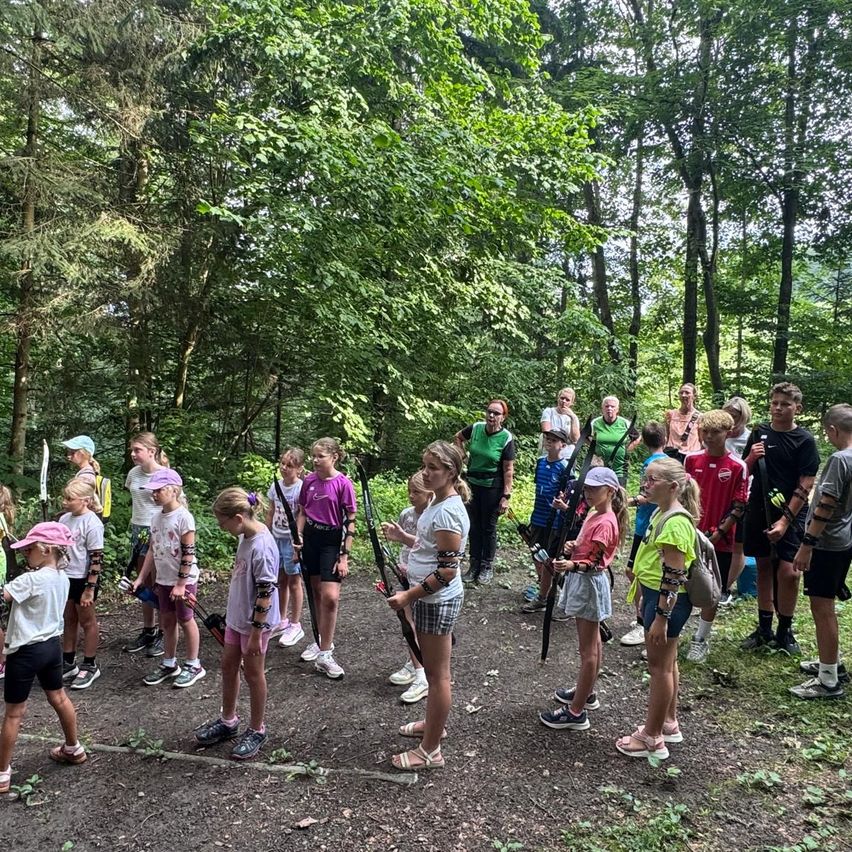 Eine Gruppe von Kindern und Erwachsenen versammelt sich in einem Wald und hält Bögen und Pfeile, wahrscheinlich an einer Bogenschießaktivität teilnehmend.