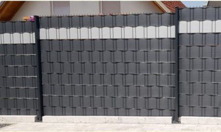 Ein moderner, großer grauer Zaun mit einem Muster kleiner Quadrate und weißen Streifen oben und unten. Er steht vor einem Haus mit einem roten Dach.