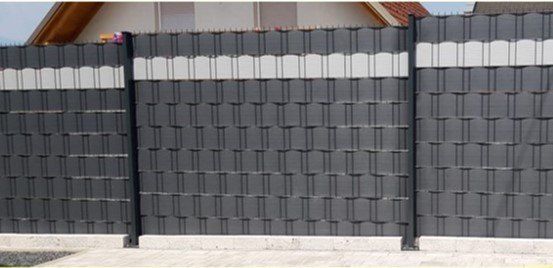 Ein moderner, großer grauer Zaun mit einem Muster kleiner Quadrate und weißen Streifen oben und unten. Er steht vor einem Haus mit einem roten Dach.
