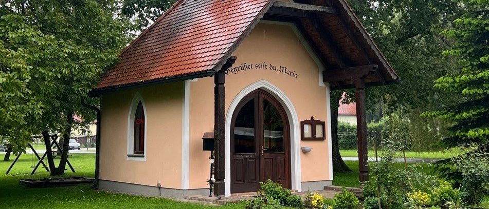 Eine kleine Kirche mit rotem Dach und Turm steht in einem Park. Grüne Bäume und Pflanzen umgeben das Gebäude. Ein Schild trägt die Aufschrift 'Geheiliget sei du, Maria.'