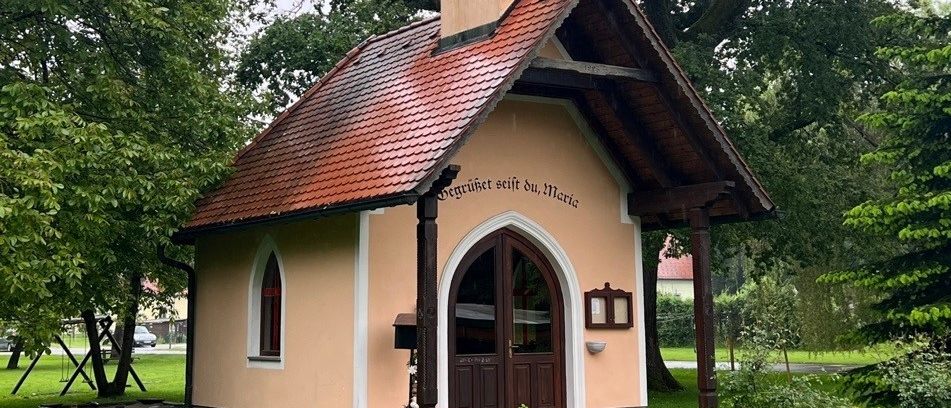 Eine kleine Kapelle mit einem Turm und einem roten Dach, umgeben von üppigem Grün und Bäumen.