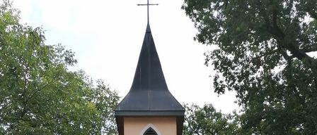 Eine kleine Kapelle mit einer deutschen Inschrift, umgeben von Blumen und Grün, hat ein schwarzes Dach und eine Holztür. Bäume und geparkte Autos sind im Hintergrund zu sehen.