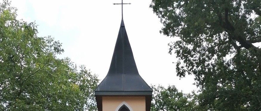 Eine kleine Kapelle mit einer deutschen Inschrift, umgeben von Blumen und Grün, hat ein schwarzes Dach und eine Holztür. Bäume und geparkte Autos sind im Hintergrund zu sehen.
