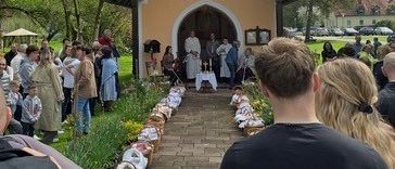 Eine kleine Kirche mit einem Turm ist zu sehen, in der eine Menschenmenge an einer Zeremonie teilnimmt. Ein Tisch mit einem Kreuz steht zwischen zwei Priestern.