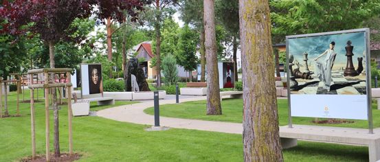 Ein gepflasterter Weg schlängelt sich durch einen Park mit grünem Gras und hohen Bäumen. Bänke, Kunstinstallationen und ein Gebäude sind in der Ferne.