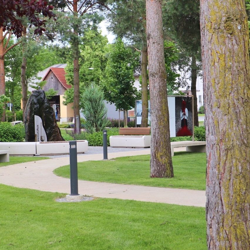 Ein gepflasterter Weg schlängelt sich durch einen Park mit grünem Gras und hohen Bäumen. Bänke, Kunstinstallationen und ein Gebäude sind in der Ferne.