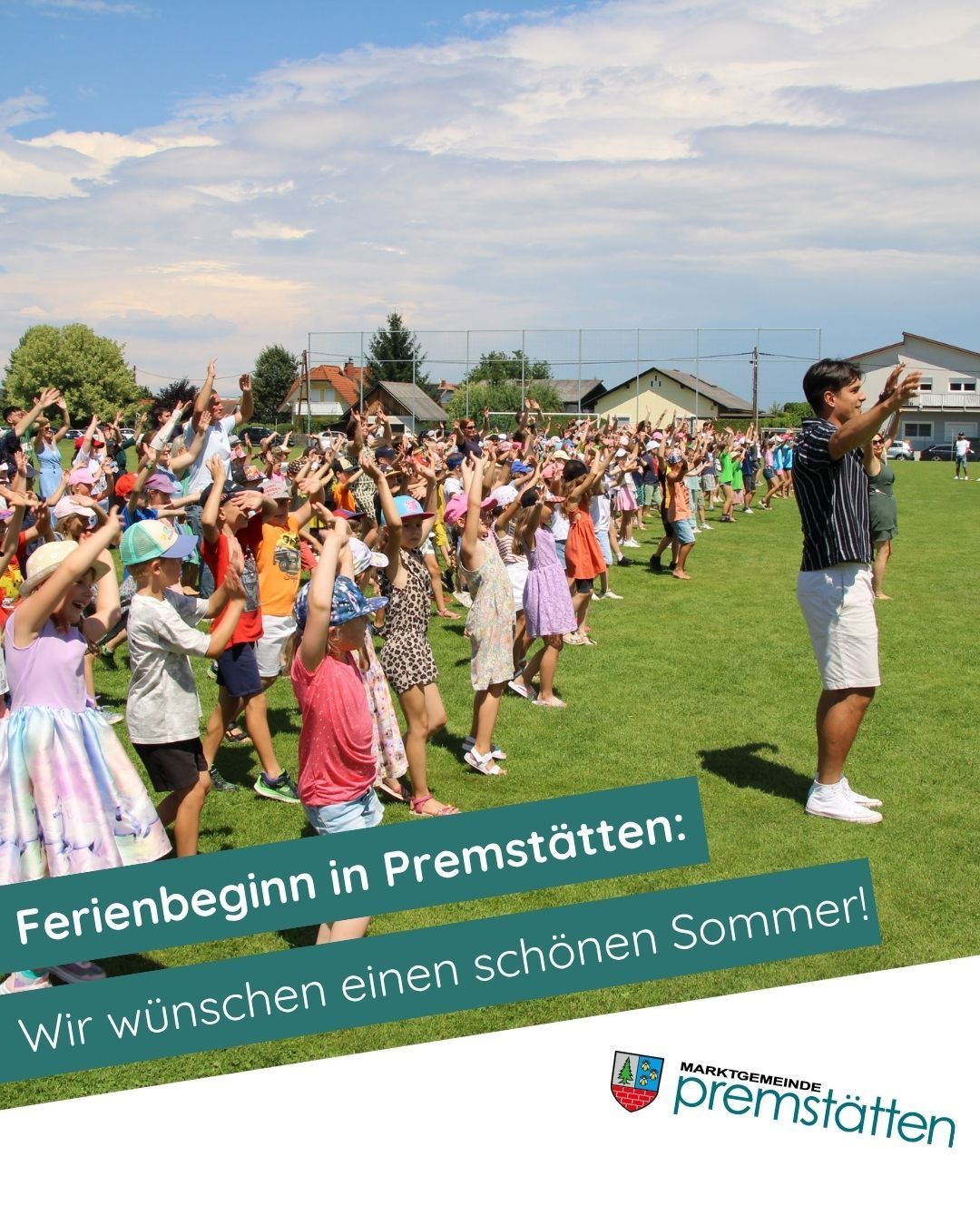 Eine Gruppe von Kindern und Erwachsenen versammelt sich auf einem Rasenfeld und hebt die Hände. Ein Mann steht vor ihnen. Sie scheinen den Beginn der Sommerferien zu feiern.