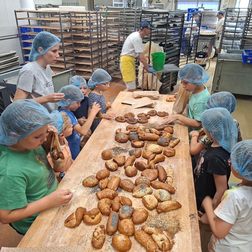 Eine Gruppe von Kindern in Haarnetzen steht um einen Tisch mit verschiedenen Broten und Bagels. Eine Frau in einem grauen T-Shirt steht neben ihnen, und ein Mann in einem weißen T-Shirt steht hinter ihnen. Es gibt Regale mit Tabletts und einem Eimer in der Nähe.