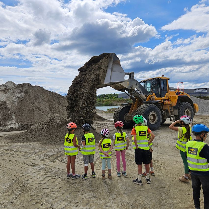 Mehrere Kinder in Warnwesten beobachten, wie ein großer Bagger Erde auf einer Baustelle abludet. Ein bewölkter Himmel ist darüber.