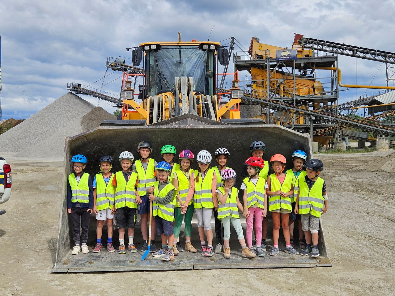 Eine Gruppe von Kindern in Sicherheitswesten und Helmen steht in einem großen Baggerlöffel, mit einem gelben Bagger im Hintergrund.