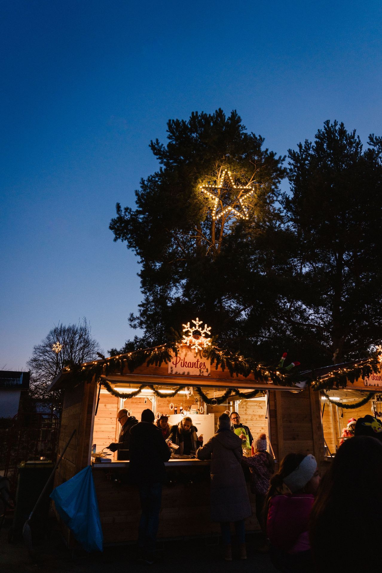 Eine Gruppe von Menschen versammelt sich bei Dämmerung um einen dekorierten Außenstand, mit einer sternförmigen Lichtinstallation darüber.