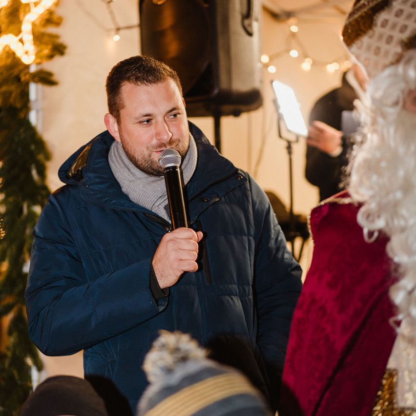 Ein Mann in einer blauen Jacke und einem Schal spricht in ein Mikrofon und sieht dabei den Weihnachtsmann in einer festlichen Umgebung.