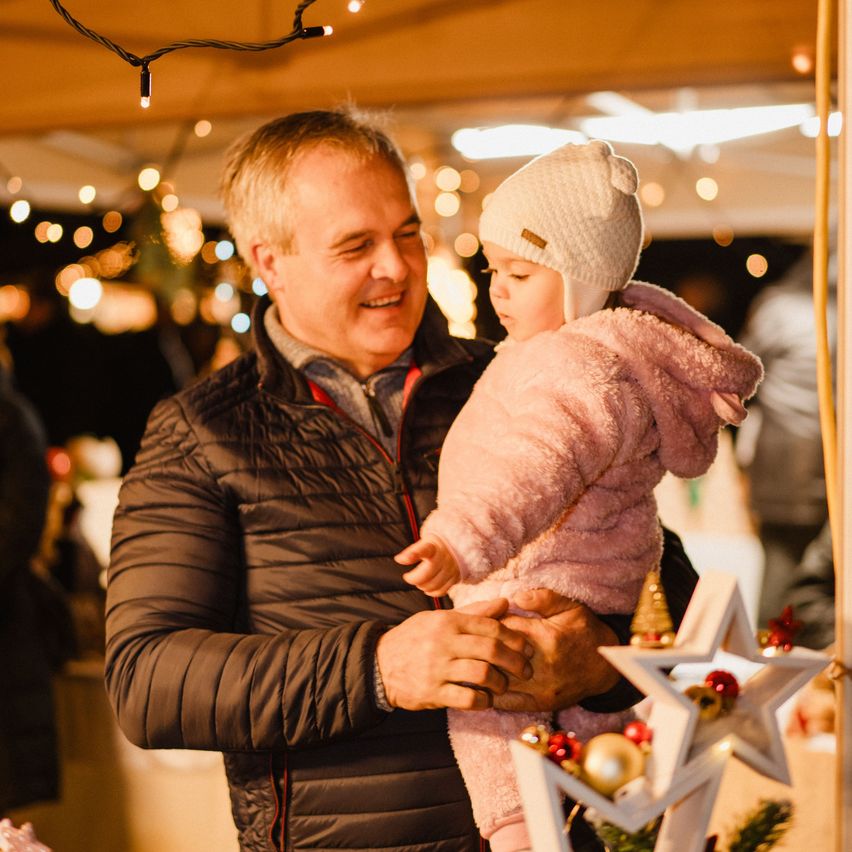 Ein Mann, der lächelt, hält ein kleines Mädchen in seinen Armen auf einem Weihnachtsmarkt, umgeben von festlichen Lichtern und Dekorationen.
