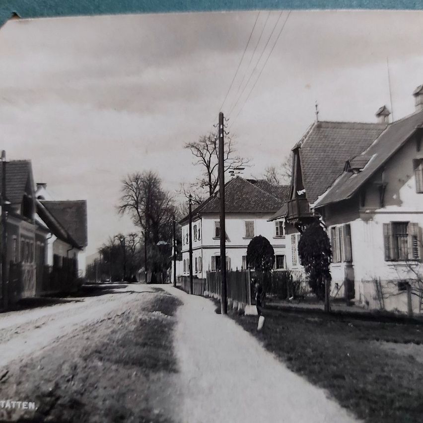 Ein Schwarz-Weiß-Foto einer alten Straße mit Häusern, Strommasten und Bäumen. Die Häuser haben Schornsteine und einige haben Fenster. Es steht eine Person am Straßenrand. Das Foto hat einen Wasserzeichen unten links.