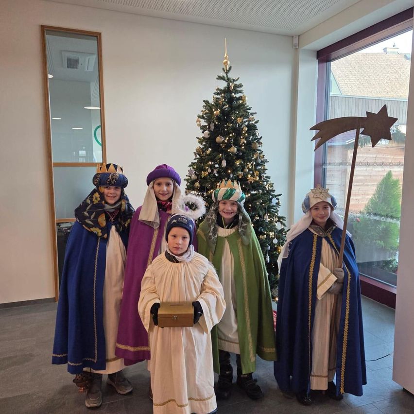 Fünf Kinder in Weihnachtskostümen stehen vor einem geschmückten Weihnachtsbaum und lächeln für ein Foto. Dahinter spiegelt ein Spiegel einen Raum mit einer Glaswand.