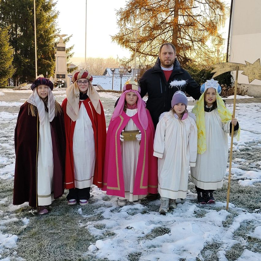 Eine Gruppe von Kindern in mittelalterlichen Kostümen steht im Schnee mit einem Mann, der einen goldenen Stab hält. Die Kinder tragen Mützen und Stiefel. Der Mann trägt eine Jacke und Hose. Hinter ihnen stehen Bäume und ein Gebäude.
