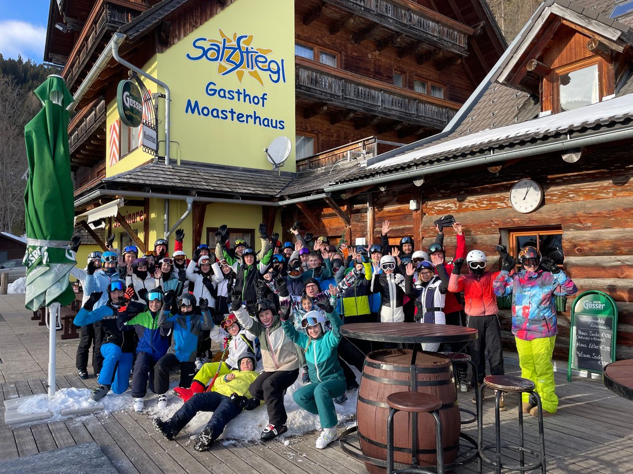 Eine Gruppe von Menschen in Wintersportausrüstung posiert für ein Foto vor einem Gebäude. Das Gebäude hat ein Schild mit der Aufschrift 'Sätzstiegel Gasthof Moasterhaus'.