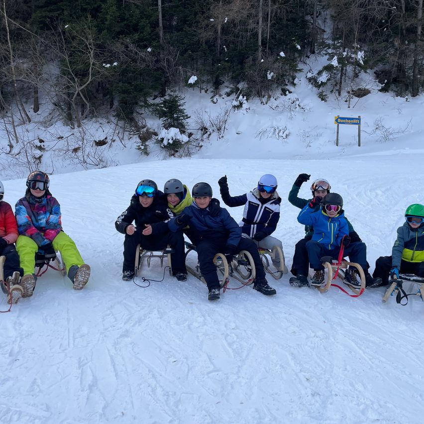 Eine Gruppe von Menschen sitzt auf Schlitten im Schnee und trägt Winterkleidung. Das Gebiet ist schneebedeckt mit einem Hintergrund aus Bäumen. Es gibt ein Schild in der Ferne.