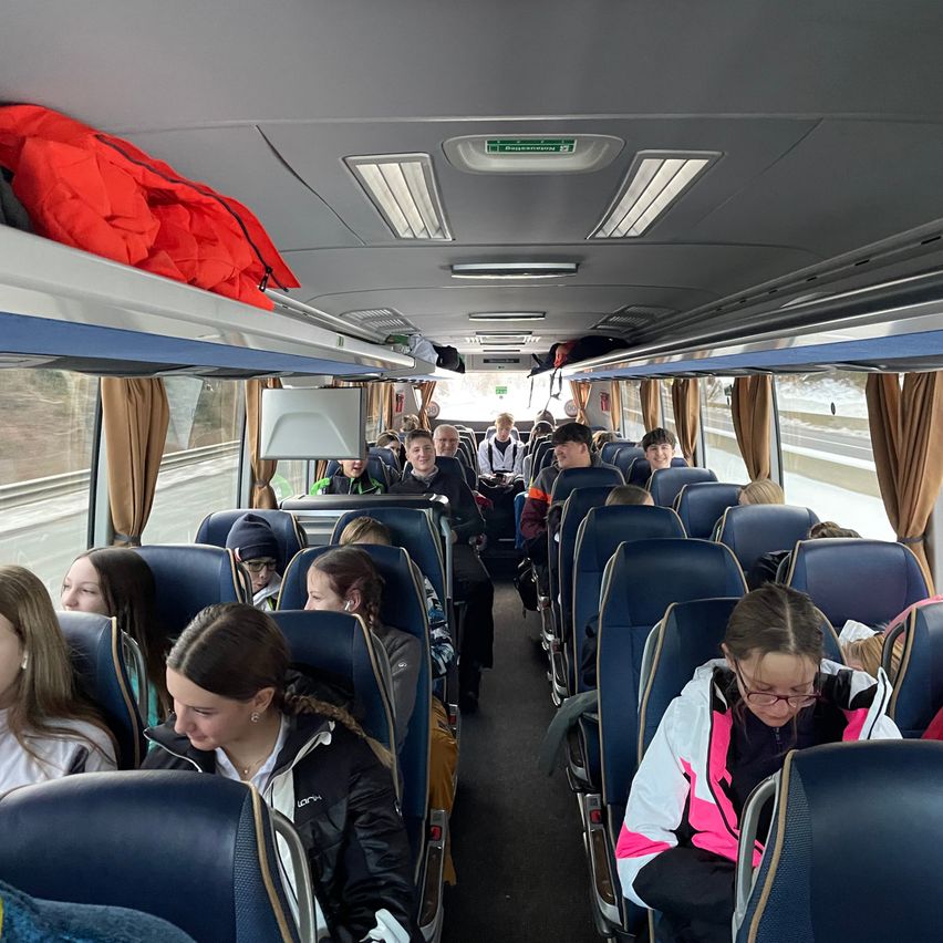 Im Inneren eines Busses sitzen mehrere Passagiere mit persönlichen Gegenständen über dem Kopf. Einige tragen Brillen, Jacken und Mützen. Eine Person steht im Gang.