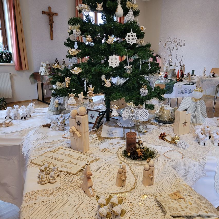 Ein Raum, der für Weihnachten geschmückt ist, mit einem Baum, Ornamenten und einem Tisch, der mit Spitze bedeckt ist. Eine Kerze steht in der Mitte des Tisches, und Figuren sind darum herum platziert. Der Raum ist mit Weihnachtsdekoration gefüllt und hat ein Holzkreuz an der Wand.