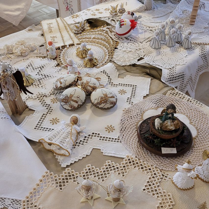 Ein Tisch ist mit verschiedenen gehäkelten Spitzen bedeckt und zeigt mehrere Engelsfiguren, eine Weihnachtskrippe und Weihnachtseier. Die Gegenstände sind festlich arrangiert.