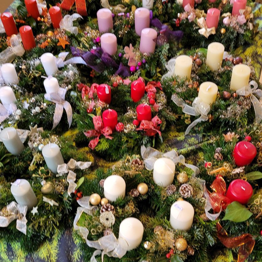 Eine Nahaufnahme von mehreren Kränzen, die in einem kreisförmigen Muster angeordnet sind und mit verschiedenfarbigen Kerzen, Bändern und Ornamenten geschmückt sind. Die Kränze sind grün mit Tannennadeln und haben verschiedene Dekorationen, darunter Sterne, Bänder und Tannenzapfen.