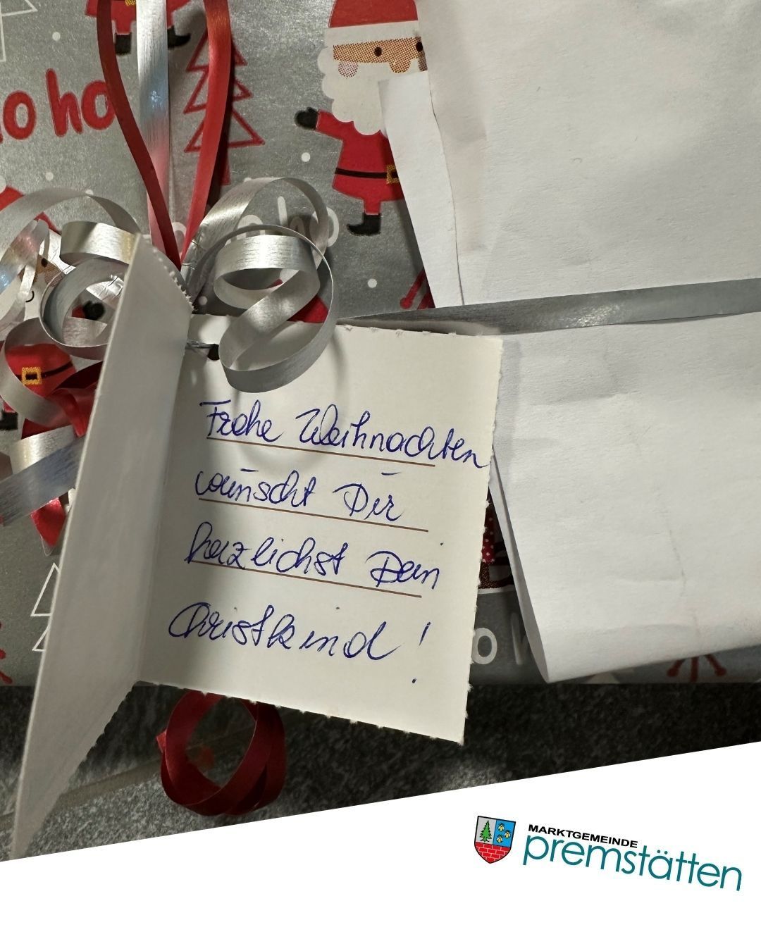Ein handgeschriebener Zettel mit blauer Tinte lautet 'Frohe Weihnachten! Gesundheit Dem Christkind!' An einem verpackten Geschenk mit silbernen Schleifen und einem roten Band befestigt. Unten befindet sich ein Logo für 'Marktgemeinde'.