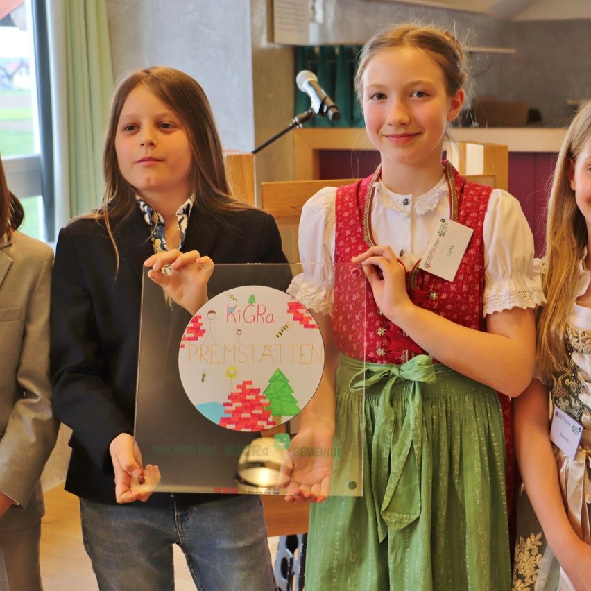 Three young girls hold a plaque with the word 'PREMSTATTEN' on it. Two of them wear traditional Bavarian clothing.