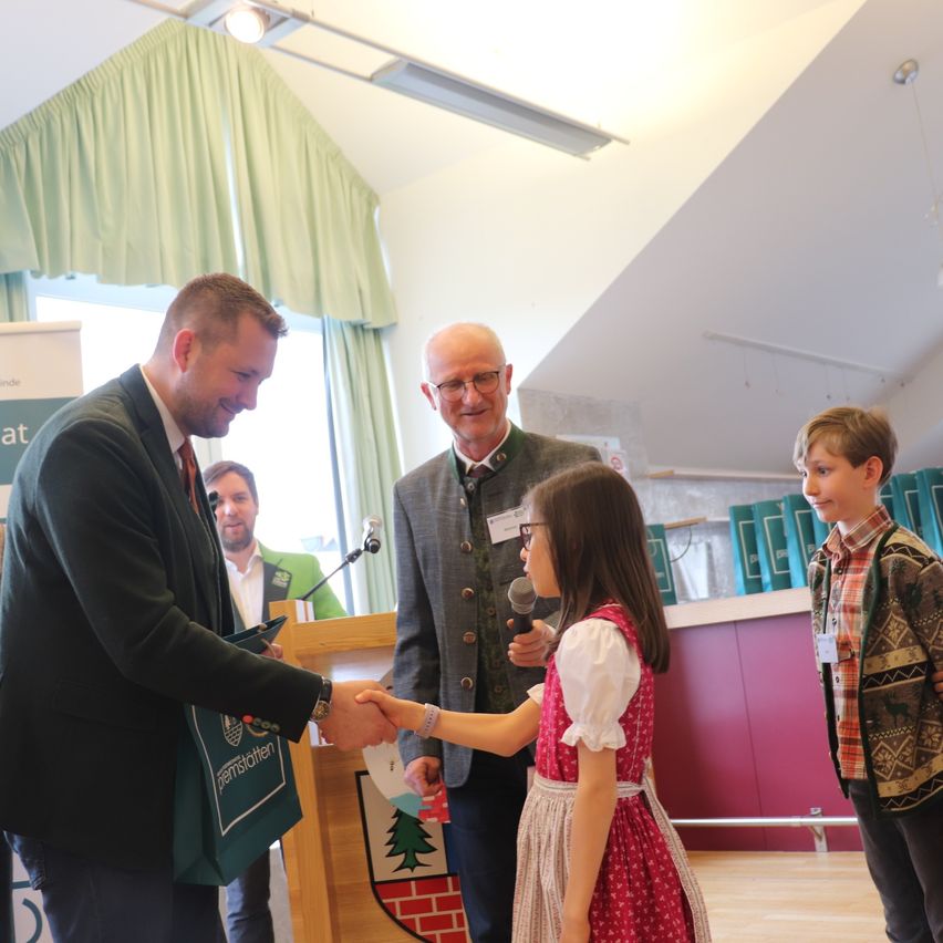 A man hands a gift to a girl in a red dress. Two boys stand behind them. One holds a microphone and speaks, while the other looks on.