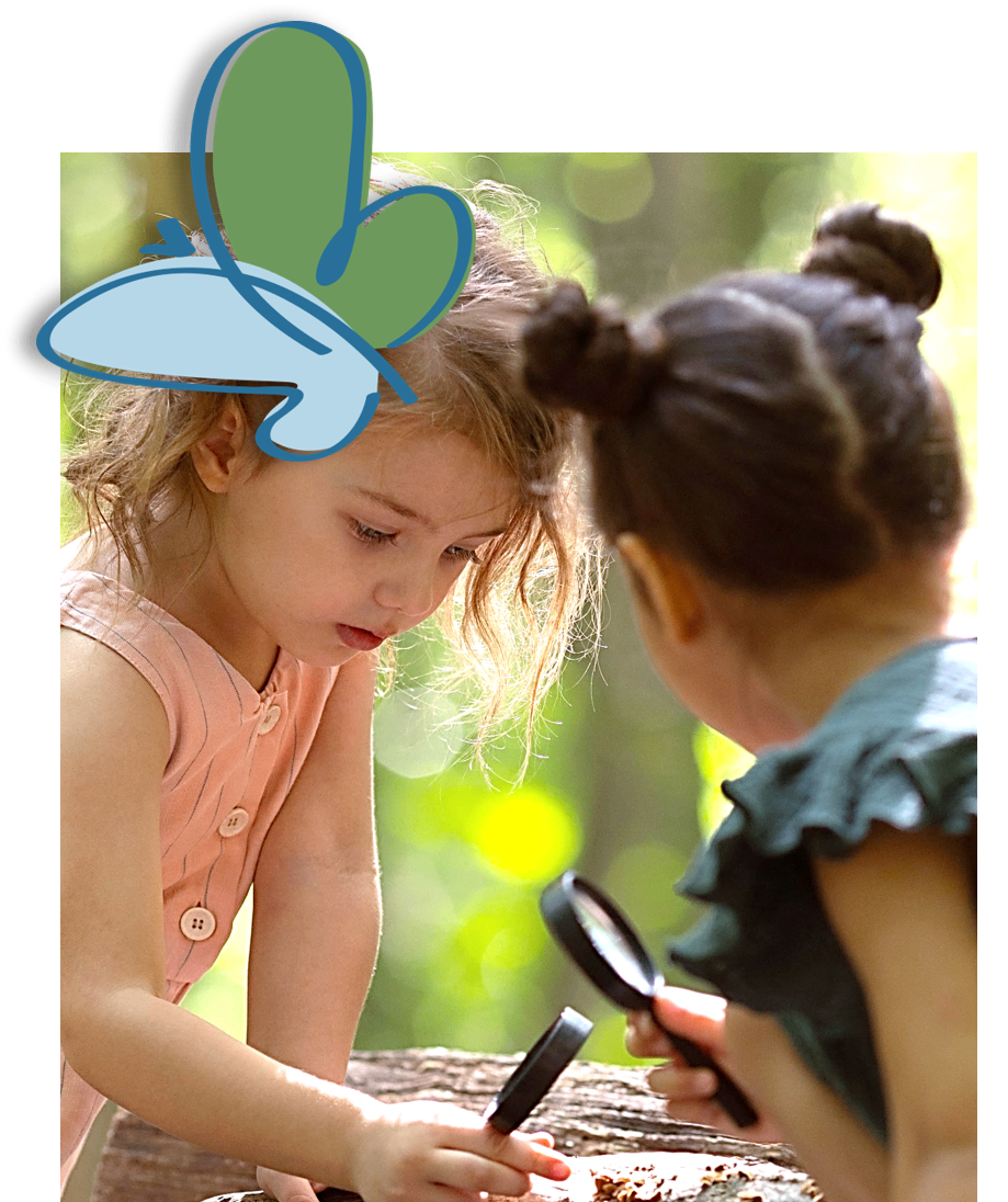 Two young girls examine something through a magnifying glass. The girl on the left has blonde hair and is wearing a pink top. The girl on the right has dark hair in a bun and is wearing a green top. They are outdoors.