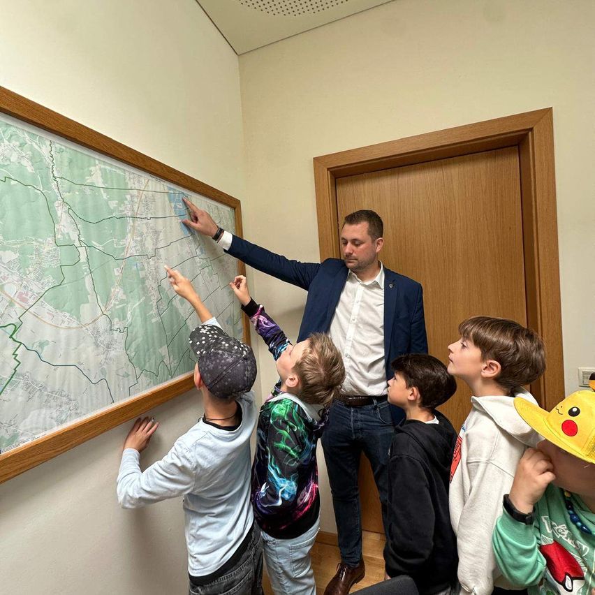 Ein Mann in einem blauen Anzug zeigt auf eine Karte in einem Klassenzimmer, während fünf Kinder zuschauen.