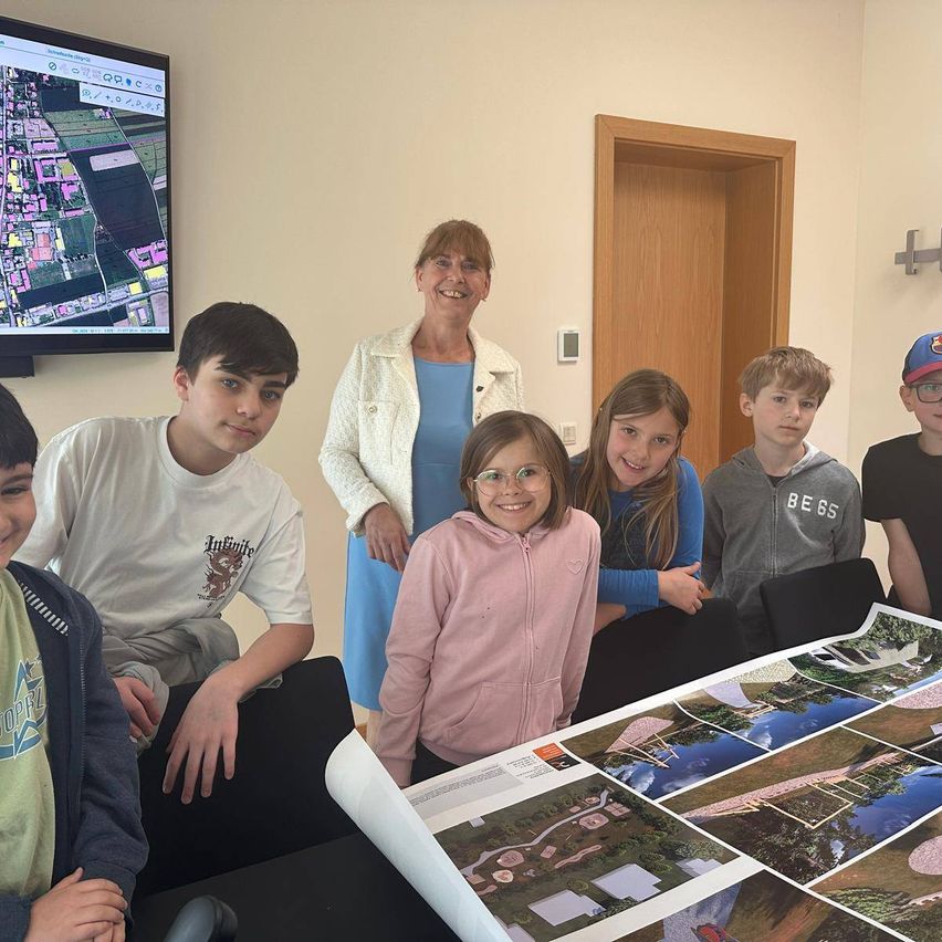Eine Gruppe von Kindern und einer erwachsenen Frau versammelt sich um einen Tisch mit Fotos und Karten. Sie scheinen über ein Projekt zu diskutieren.