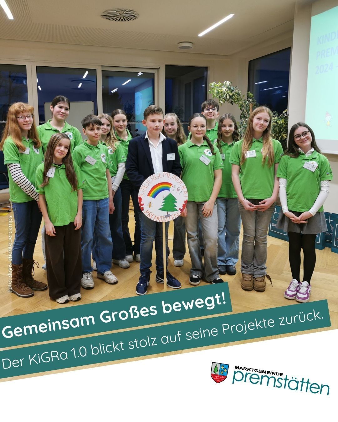 Eine Gruppe von Kindern in grünen Shirts steht vor einem Regenbogenschild, mit einem Mann im Anzug in der Mitte, der auf eine Projektionsleinwand schaut.