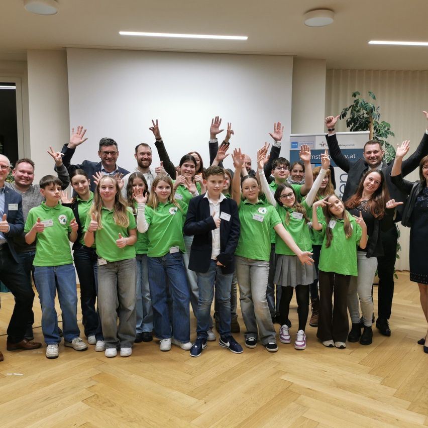 Eine Gruppe von Menschen in grünen Shirts und Jeans posiert für ein Foto mit erhobenen Händen und Lächeln in einem Raum mit Holzboden und weißer Wand.
