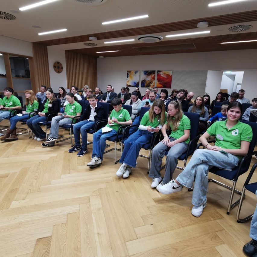 Eine Gruppe von Personen in grünen Shirts sitzt in einem Raum mit Holzboden und weißen Wänden. Sie lauschen aufmerksam jemandem vorne, einige halten Papiere und andere sind mit ihren Telefonen beschäftigt.