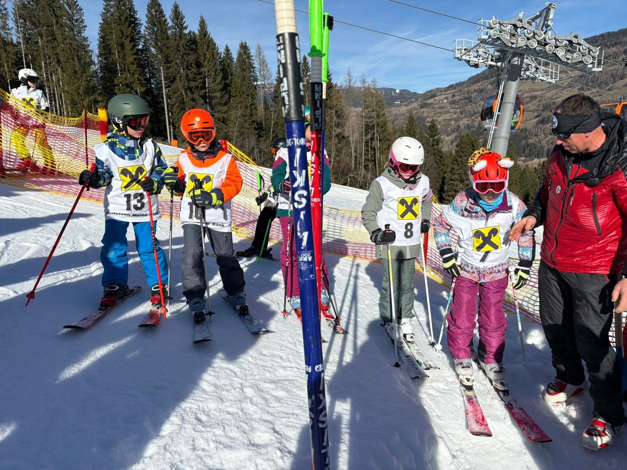 Eine Gruppe von Kindern in Skiausrüstung und Helmen steht in einem verschneiten Bereich, wahrscheinlich in einem Skigebiet. Sie sind mit den Nummern 13, 10, 8 und 7 versehen.