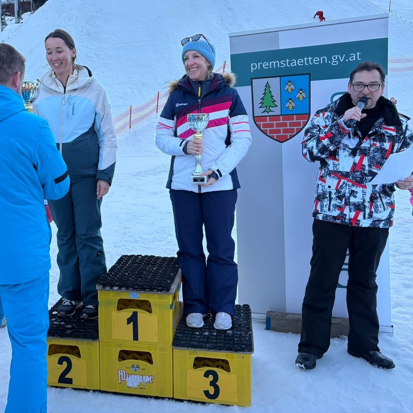 Drei Personen stehen auf einem verschneiten Podest mit den Nummern 1, 2 und 3. Eine Frau in der Mitte hält einen Pokal. Eine weitere Frau und ein Mann, beide mit Mikrofonen, stehen in der Nähe. Hinter ihnen ist ein Schild mit einem Logo und dem Text 'premstaetten.gv.at'.