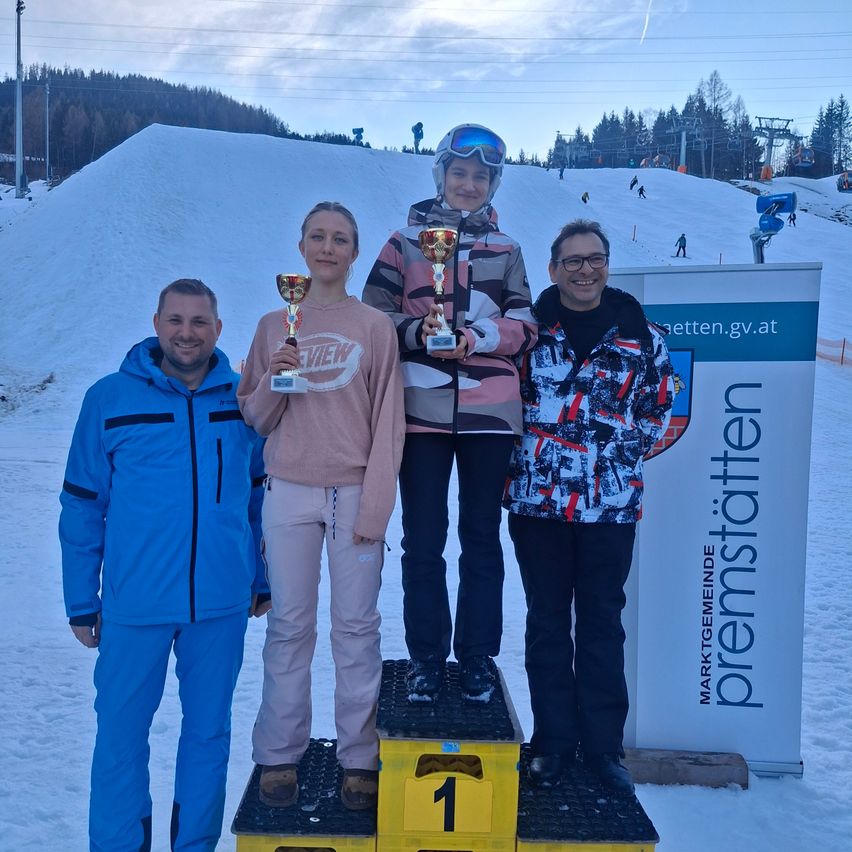 Vier Personen stehen auf einem Podest in einem Skigebiet. Zwei Frauen halten Pokale und zwei Männer stehen neben ihnen. Dahinter befindet sich ein verschneiter Hügel und ein Schild mit dem Text 'premierstatten'.