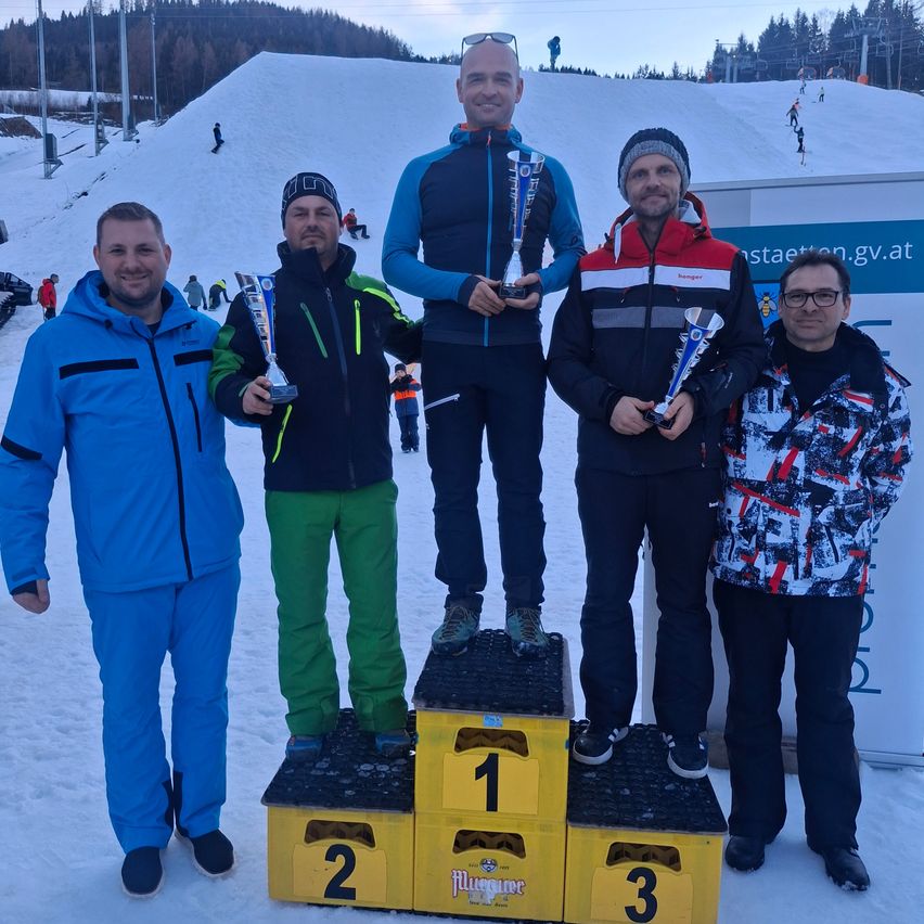 Fünf Männer stehen auf einem verschneiten Podest, tragen Winterjacken und halten Pokale. Der Sieger des ersten Platzes trägt Blau, der Zweite trägt Grün. Der Dritte trägt Rot. Im Hintergrund sind verschneite Hänge und Skifahrer zu sehen.