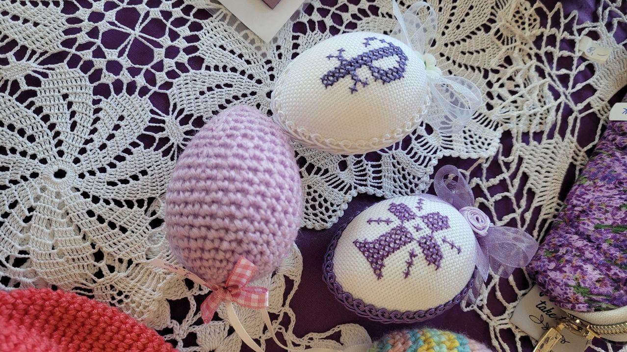 Drei dekorative Ostereier auf einer Spitzendeckchen. Ein Ei ist in Lila gehäkelt, ein weiteres mit weißen Stickereien und lila Designs bestickt und das dritte ist eine Mischung aus Häkeln und Sticken mit lila Designs.
