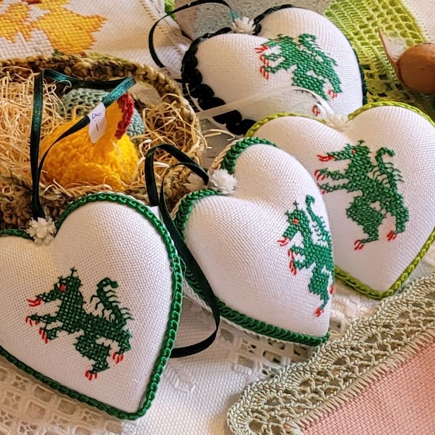 Mehrere herzförmige gestickte Kissen mit grüner Drachenstickerei sind auf einem weißen Tuch neben einem Korb mit einem gestrickten Huhn und anderen Gegenständen ausgestellt.