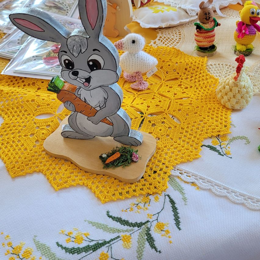 Eine hölzerne Hasenfigur mit einer Karotte steht auf einem gelben Doily mit Blumenmustern. Andere Figuren und Gegenstände sind auf dem Tisch.